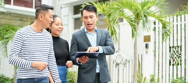 Asian young realtor showing photos of new house to young family on tablet pc while they walking along the street