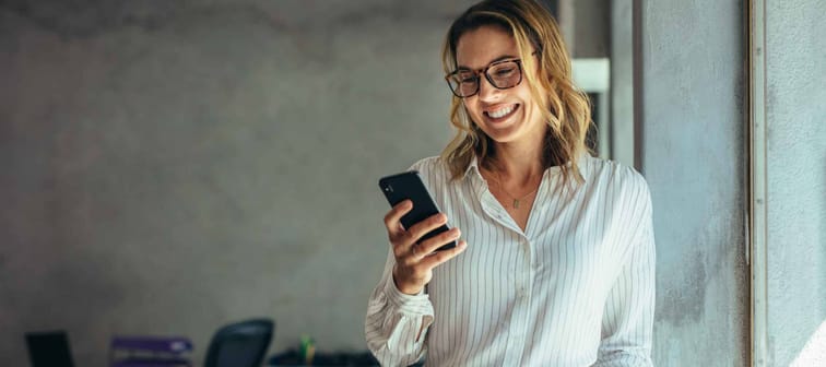 Smiling businesswoman using phone in office. Small business entrepreneur looking at her mobile phone and smiling.