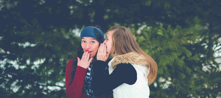 two women in winter in forest. One whispering investment tips in the other's ear. The other is excited by the news.