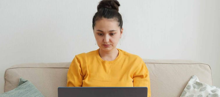 Woman blogger influencer working on the computer at home on the couch. Young asian woman in yellow sweater with focused gaze working remotely at home during pandemic