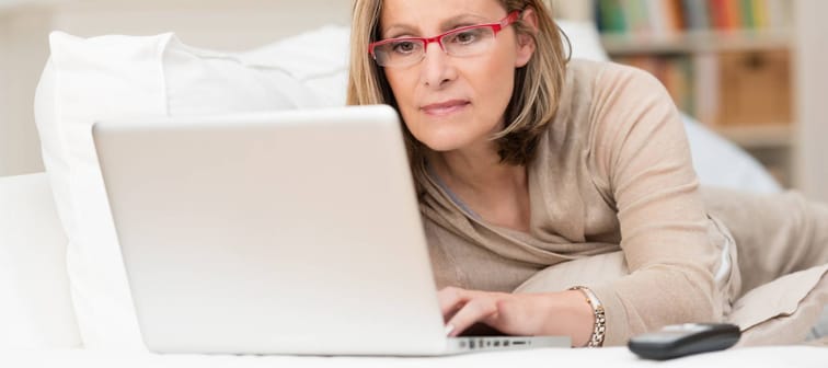 Woman wearing glasses lying on a sofa at home concentrating as she works on a laptop