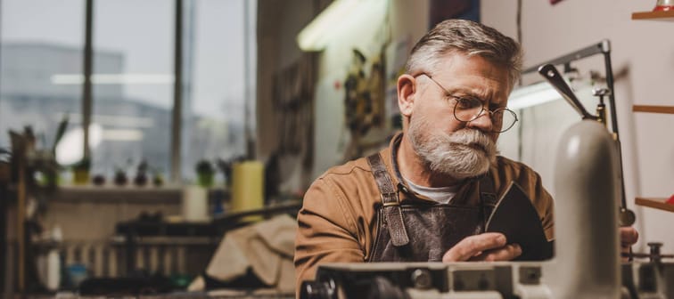 Senior, bearded cobbler sewing leather on sewing machine in workshop